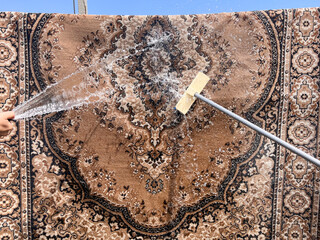 A carpet being washed outdoors using a hose and brush, water flowing over the surface, showing the cleaning process in progress under natural light.