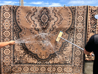 A traditional carpet being washed outdoors with a hose and scrub brush. A woman's hand and a man's hand are seen from opposite sides, actively cleaning the rug with water.
