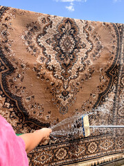 A vertical shot of a woman’s hand in a pink t‑shirt holding a garden hose spraying water onto a carpet being washed outdoors.