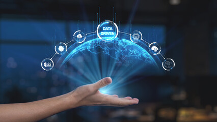 A hand is reaching out towards a glowing globe, illustrating a digital network of icons that symbolize data analytics and technological advancement in a modern workspace. Synapse