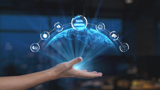 A hand is reaching out towards a glowing globe, illustrating a digital network of icons that symbolize data analytics and technological advancement in a modern workspace. Synapse