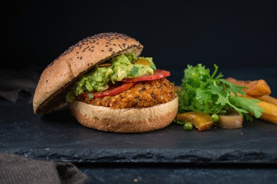 Nutritious roasted sweet potato patty with whole wheat bun, avocado spread, plant-based mayo, and fresh veggies on a dark surface. Vegan meal idea, moody food photography. - Powered by Adobe