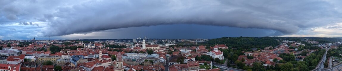 Wide panoramic view of Vilnius, Lithuania, with red rooftops and historic architecture beneath a dark storm front and dramatic sky.