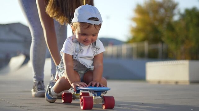 Toddler on skateboard with parent guiding child and baby at skatepark mother supporting playful child as toddler smiles while playing outdoors on concrete child enjoying outdoor play and skateboard