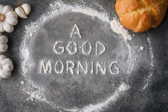 Morning greetings spelled out with baking flour next to freshly baked spiced bread and garlic on a neutral surface. Image for culinary classes. Homemade treats. Home cooking. High resolution photo. - Powered by Adobe