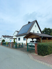 Naklejka premium Solar panels gleam on a modern home in a peaceful neighborhood on a cloudy day with a hint of greenery and paved walkways surrounding the property