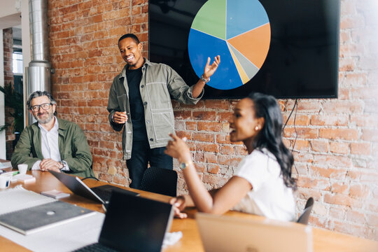 Happy male business professional pitching while explaining pie chart to colleagues in office