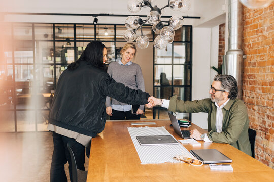 Happy woman shaking hands with male recruiter sitting at table for job interview in office