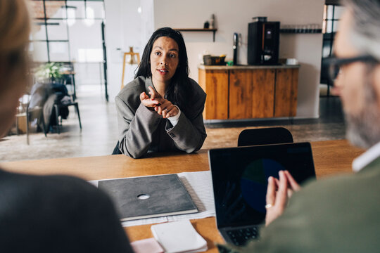Woman gesturing while giving interview to business experts while sitting at table in office