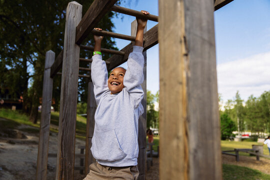 Happy boy hanging on monkey bar near forest during summer camp