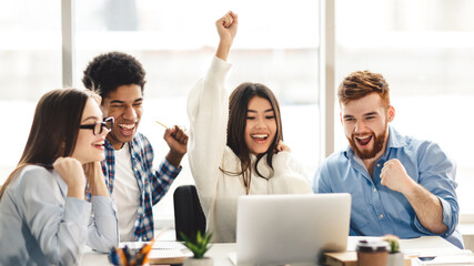 A group of multiethnic students joyfully sit around a desk, raising their arms in celebration....