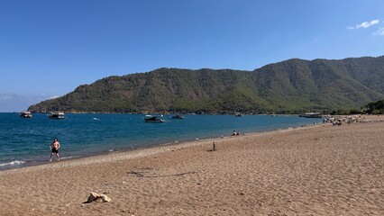 A wide sand and pebble beach with vacationers and moored tourist boats against the backdrop of forested mountains under the bright midday sun. Port of Adrasan, Turkey.