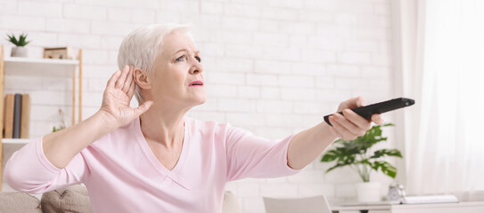 Elderly woman making hearing sign, rising tv set volume with remote control, panorama
