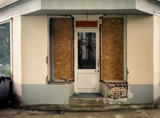 Closed and abandoned storefront with boarded-up windows and damaged facade. The old shop entrance shows signs of decay, cracked plaster, exposed bricks, and a neglected exterior.