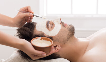 Unshaven man getting facial nourishing mask by beautician at spa salon