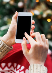 Christmas online shopping. Female buyer makes order at screen of smartphone with copy space. Woman buys presents for xmas eve, sits at home near decorated xmas tree. Winter holidays sales