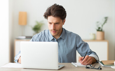 Budget Planning. Serious Man Working On Laptop And Taking Notes Indoor.