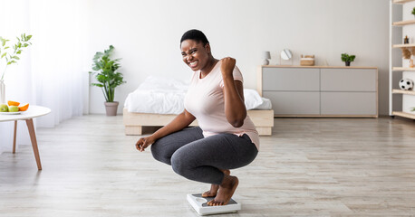 Obraz premium Excited curvy black woman sitting on scales at home, making YES gesture, happy with result of her slimming diet, panorama. Overjoyed plus size African American lady achieving her weight loss goal