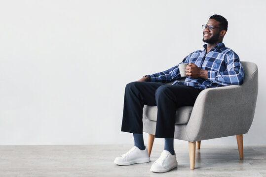 Coffee break. Dreamy young African American man sitting in armchair, enjoying cup of hot beverage against white studio wall, copy space. Millennial black guy relaxing with mug of warm drink