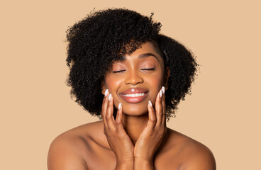 Tranquil young black woman with eyes closed, hands gently caressing her cheeks, exuding peace and serenity posing against soft beige background