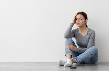 Frustrated sad worried caucasian young woman waiting for call and thinking, sitting on floor at home, on gray wall background. Nervous stress, depression and bad news and covid-19 outbreak, copy space