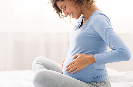 Happy expectant woman touching belly, enjoying unborn baby pushings in bed, side view
