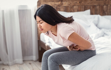 Asian Girl Having Abdominal Pain Touching Aching Stomach Sitting In Bed At Home. Intestinal Issues Concept