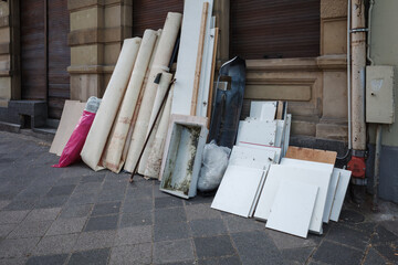 A pile of bulky waste, known as Sperrmüll in Germany, sits on a city sidewalk in Berlin awaiting collection. The discarded items include old rolled-up carpets, disassembled white cabinet parts.