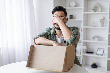 Wrong Item. Upset Arab Guy Opening Parcel And Making Facepalm Gesture While Sitting At Table At Home, Young Middle Eastern Man Feeling Sad After Unpacking Box With Delivery, Copy Space © Prostock-studio