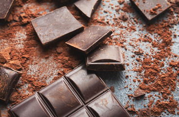 Dark chocolate bar broken in pieces on dark background and cocoa powder closeup, copy space. Sweet wallpaper, confectionery shop advertising and cooking ingredients concept