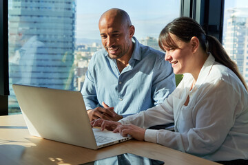 Fototapeta premium Two multiracial business colleagues sitting in the office working using a laptop discussing a project strategy together with the city in the background. Image with copy space.