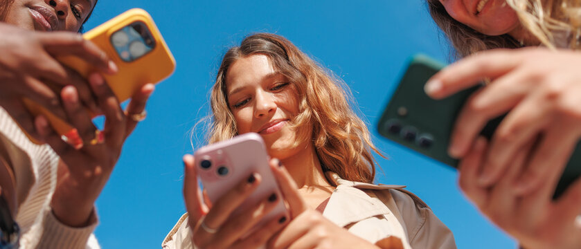 Diverse women holding smartphones, engaging in social media and digital connection under a blue sky - Powered by Adobe