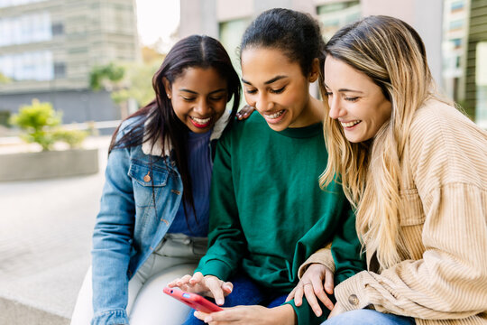Multiracial group of three young women is sitting close on a bench at university campus, using a mobile phone together and sharing content. Student lifestyle, social media, youth connection concept