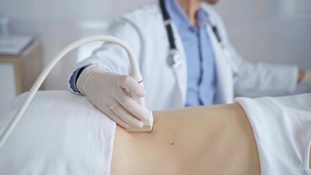 Doctor man wearing white protective gloves is using ultrasound equipment on the back of a patient, woman. Physician analyzing the results on a monitor in the background. Medicine - Powered by Adobe