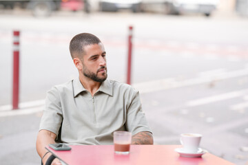 Fototapeta premium A man sits at a table with a cup of coffee and a cell phone. He is looking at the phone, possibly checking messages or browsing the internet. Concept of solitude and contemplation