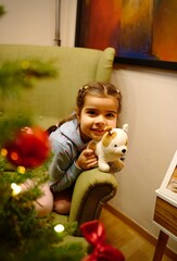 Cute little girl peeking from behind a decorated Christmas tree at home. Festive holiday atmosphere with lights, ornaments, and joyful winter mood. Perfect image for Christmas, New Year