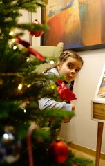Cute little girl peeking from behind a decorated Christmas tree at home. Festive holiday atmosphere with lights, ornaments, and joyful winter mood. Perfect image for Christmas, New Year