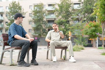 Two men sitting on a bench in a park. One of them is talking on his cell phone