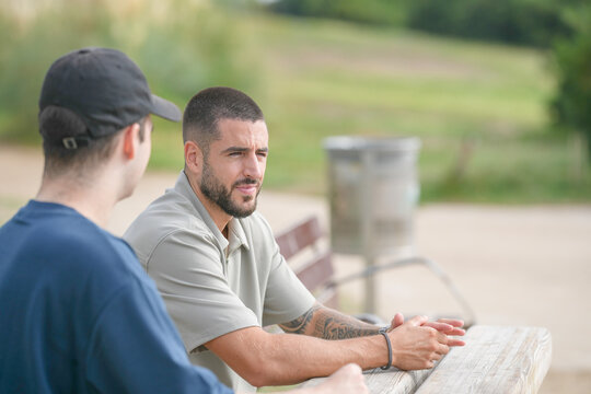 Two men are sitting on a bench in a park. One of them is wearing a hat and the other is wearing a blue shirt. They are talking to each other