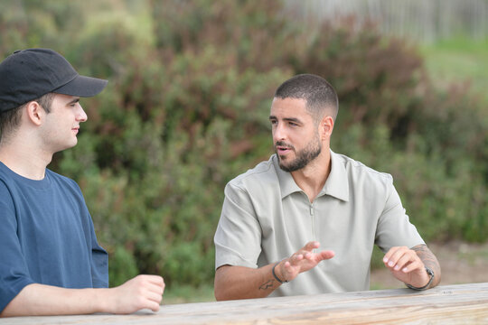 Two men are sitting at a picnic table, one wearing a black hat. They are talking to each other - Powered by Adobe