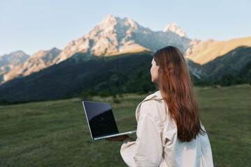 A woman stands in a grassy field with a laptop, facing distant mountains under clear skies. She...