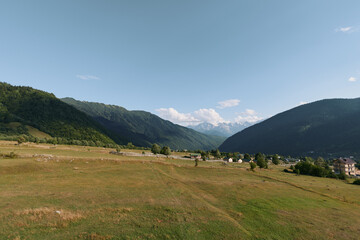 A tranquil rural landscape features open meadows stretching toward distant hills under a clear blue sky. Soft sunlight highlights grass and scattered trees in a peaceful setting.
