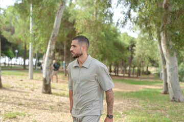 Obraz premium A man in a tan shirt walks through a park. The park is full of trees and grass, and there are a few other people in the background. The man is alone, and he is wearing a watch