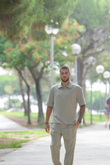 Obraz premium A man in a tan shirt and khaki pants walks down a sidewalk. The man is wearing a watch and has a beard. The scene is peaceful and calm, with trees in the background