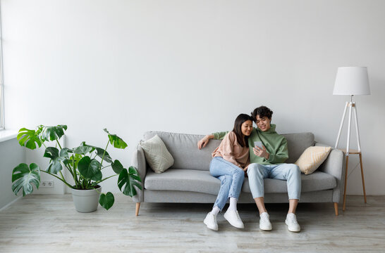 Happy millennial Asian couple using cellphone, texting and browsing internet, relaxing on couch indoors, copy space. Relaxed young family using mobile application, resting at home