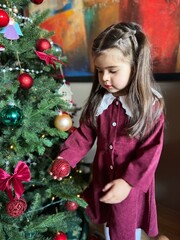 Cute little girl staying near a decorated Christmas tree at home. Festive holiday atmosphere with lights, ornaments, and joyful winter mood. Perfect image for Christmas, New Year