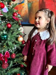 Cute little girl staying near a decorated Christmas tree at home. Festive holiday atmosphere with lights, ornaments, and joyful winter mood. Perfect image for Christmas, New Year
