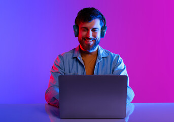 Freelance And Gaming. Happy Millennial Guy Wearing Headphones Browsing Internet On Laptop Computer, Sitting At Desk While Working Or Playing Online Game Over Neon Purple Blue Studio Background © Prostock-studio