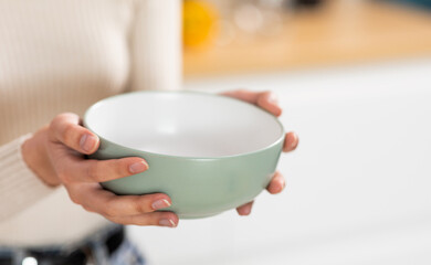 Unrecognizable woman holding empty bowl in her hands, kitchen interior, copy space, closeup shot, cropped of hungry lady starving. Famine, poverty, hunger, crisis concept