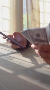 Close-up of man's hand holding smartphone and hundred dollar bills while taking notes, money financial management concept. Soft focus, Selective focus